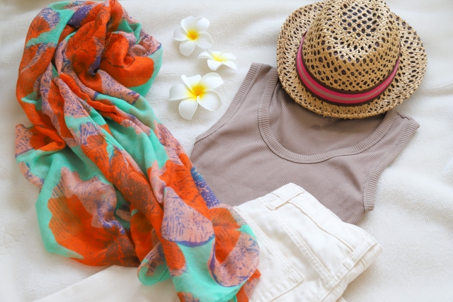 白い布の上に置かれた麦わら帽子とベージュのタンクトップと白いパンツとカラフルなスカーフとプルメリアの花